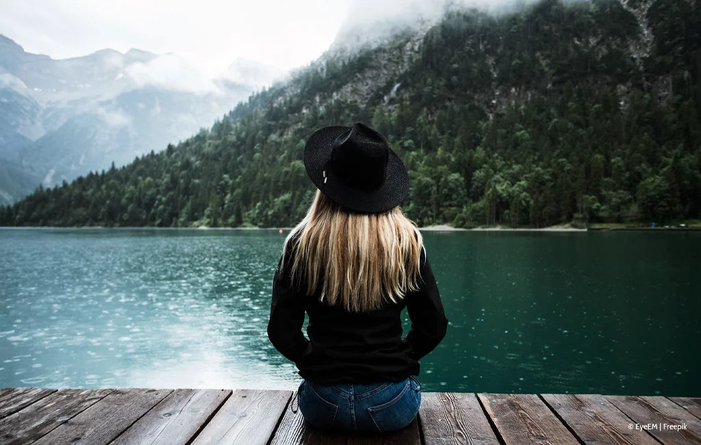 Das Bild zeigt eine Frau, die entspannt auf einem Steg sitzt, umgeben von einer malerischen Tiroler Landschaft mit Bergen und Wasser. Die Szene strahlt Ruhe, Naturverbundenheit und Gelassenheit aus.
