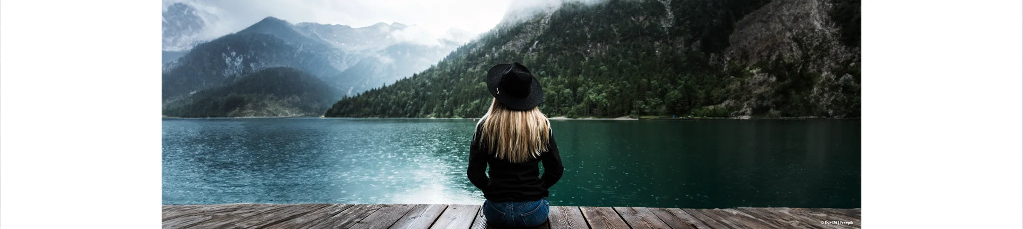 Das Bild zeigt eine Frau, die entspannt auf einem Steg sitzt, umgeben von einer beeindruckenden Berglandschaft. Die Szene strahlt Ruhe, Naturnähe und Gelassenheit aus.